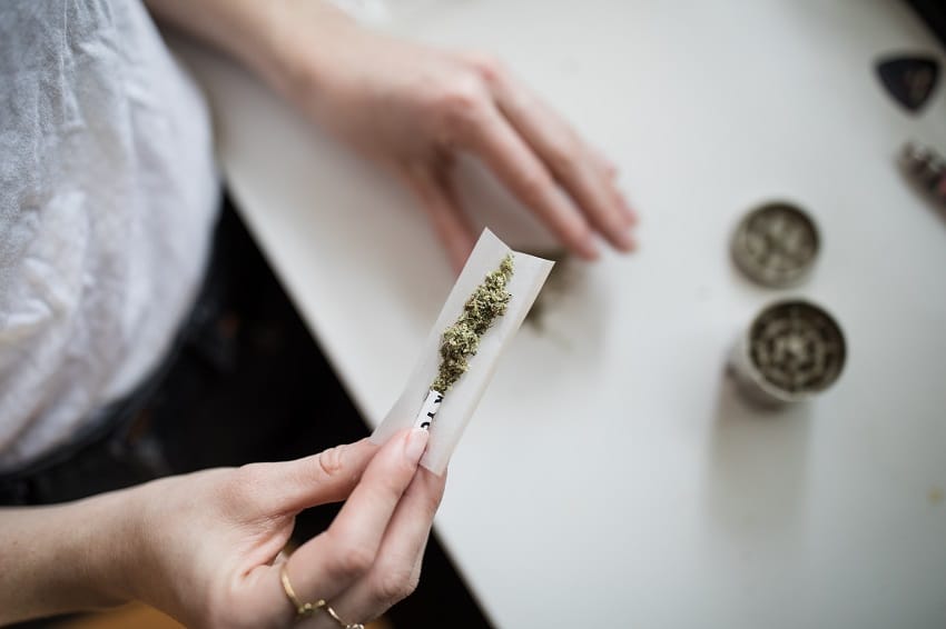 Hand holds rolling paper to roll joint with freshly-ground herb for smoking beside metal grinder on countertop