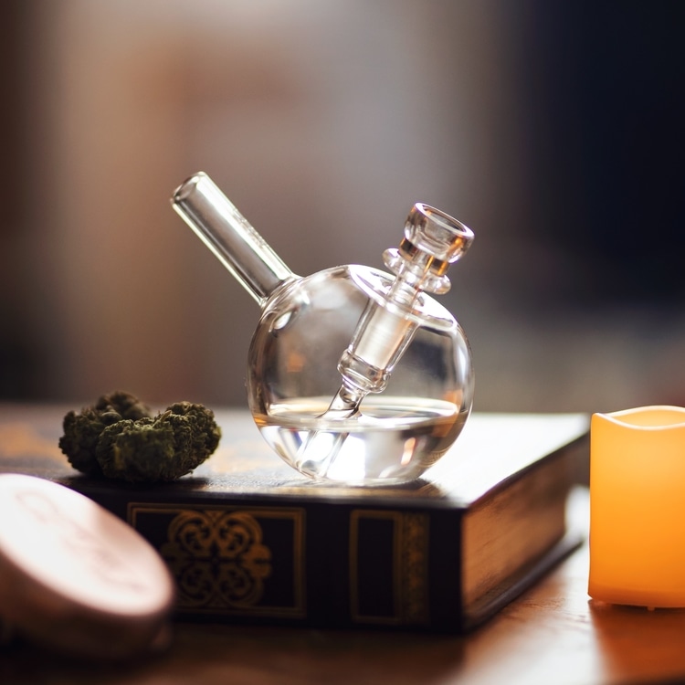 Translucent water pipe and cannabis nugget on research book beside candle