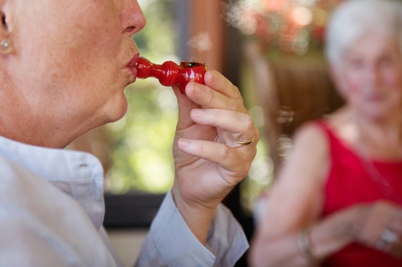 Elderly woman holding red hand pipe to lips beside friend