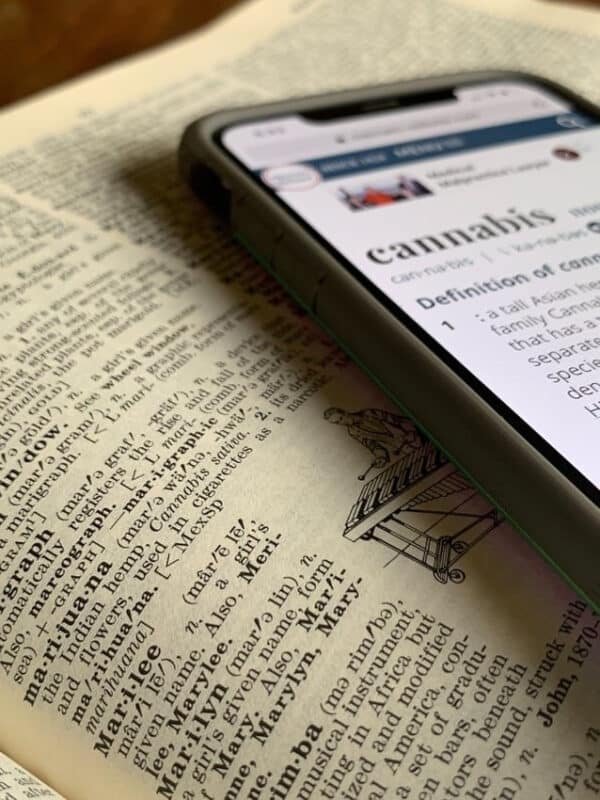Phone displaying online dictionary definition of "cannabis," resting on paper dictionary turned to definition of "marijuana"