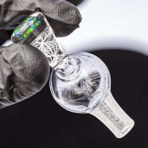 Mycomann engraved glass bubble cap with opal handle - held between two fingers of glass gloved hand.