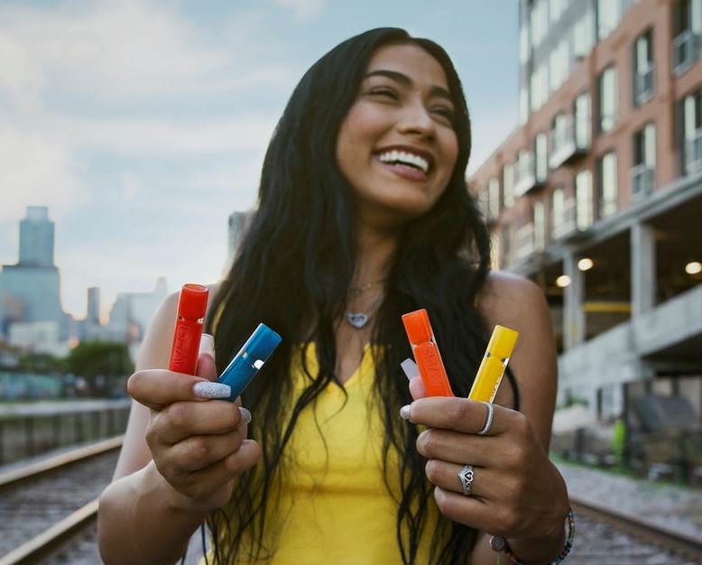 Smiling Woman Holding Chillums in Red, Blue, Orange and Yellow Outdoors on Cloudy Day