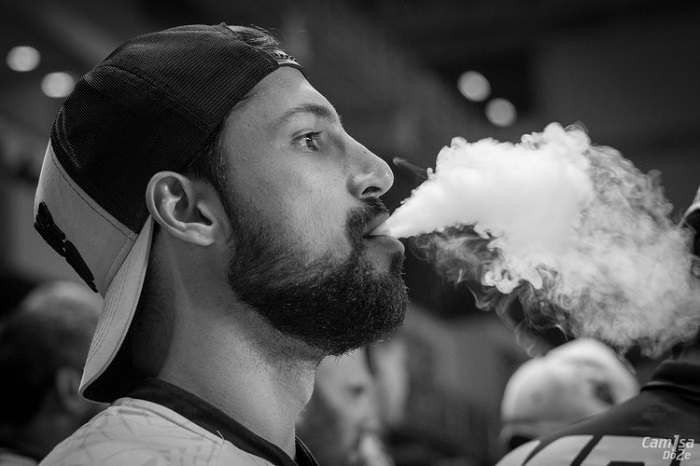 Man in baseball cap exhaling vape cloud while using dry herb vaporizer