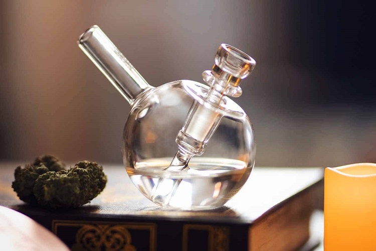 Transparent bong containing clean bong water on hardcover book beside cannabis nugget and candle