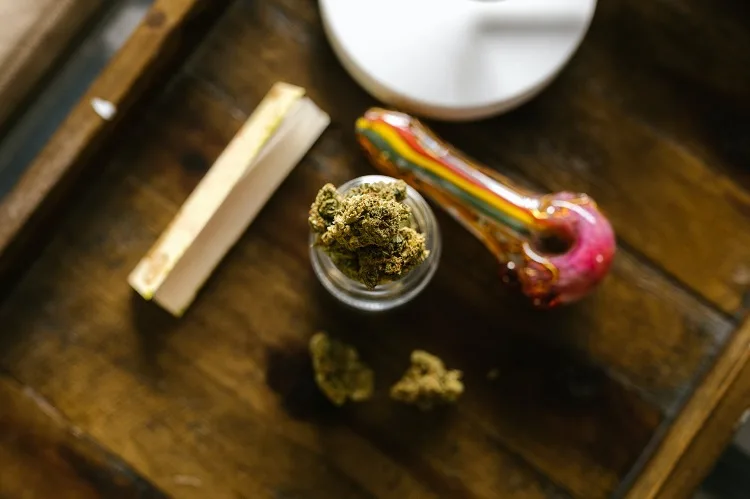 Rolling paper, glass spoon pipe and cannabis nuggets in miniature jar on wooden rolling tray