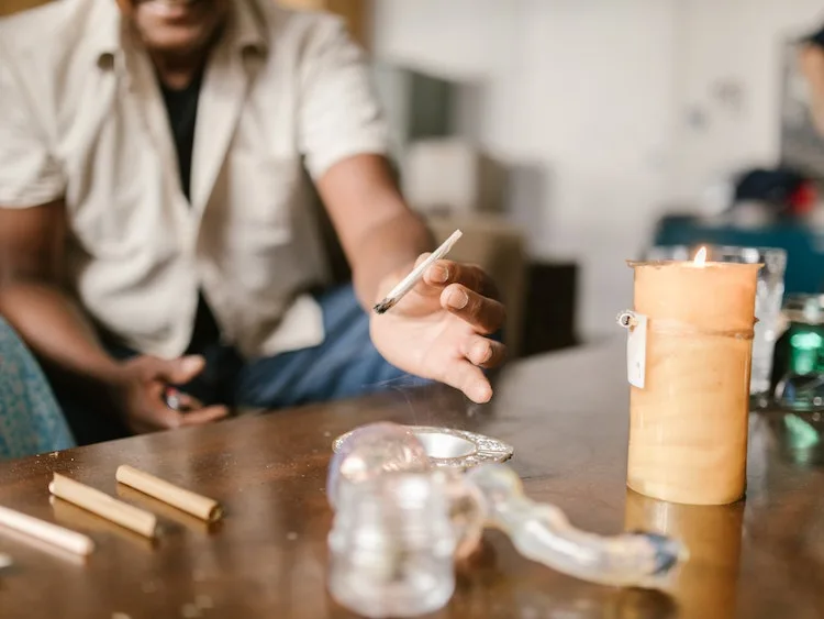 Man on couch holding lit joint above table with glass pipe and candle