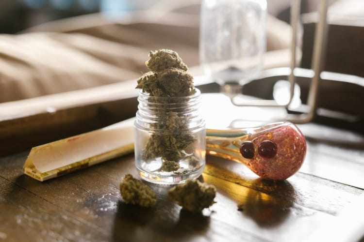 Pink and yellow spoon pipe, jar with cannabis nuggets and RAW brand rolling papers on wooden bedside table.