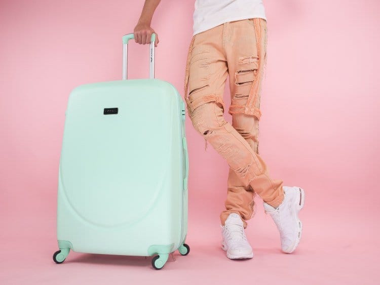 Man standing with crossed leg beside light turquoise suitcase in pastel pink room