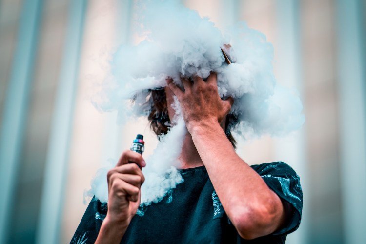Long-haired man exhaling large vaporizer cloud while partially covering mouth with hand