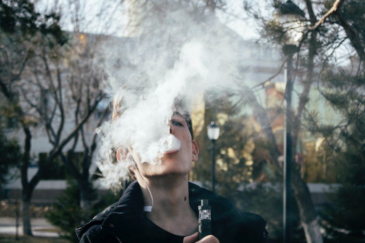 Short-haired man holding vaporizer and exhaling vape cloud in outdoor plaza