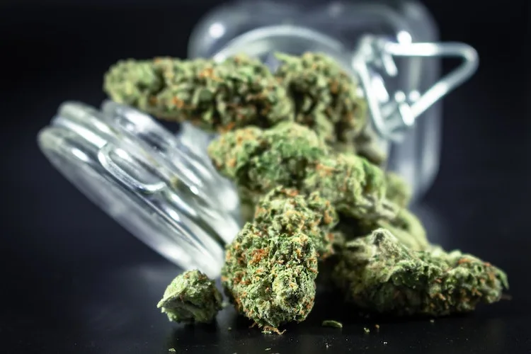 Cannabis nuggets spilling from clear glass storage container on black surface