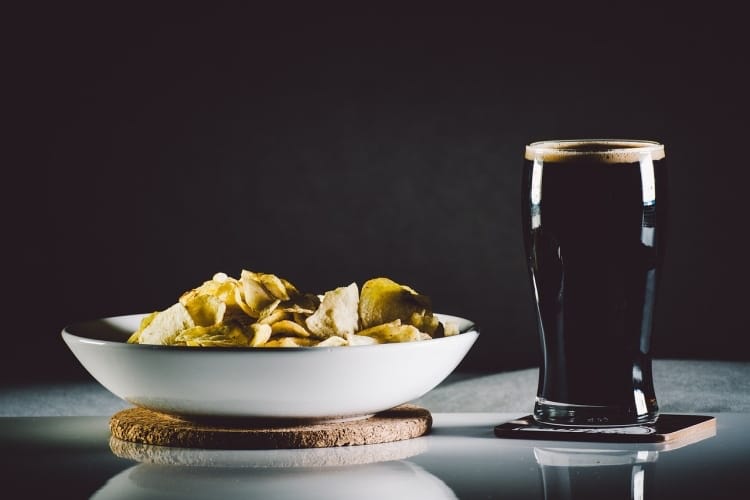 White bowl of potato chips beside porter beer in pint glass on gray table