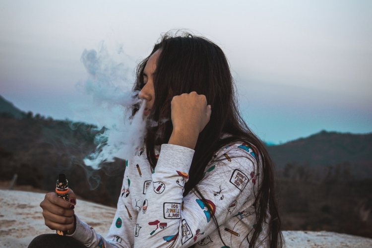 Person exhaling vape cloud through nose while holding vaporizer outdoors