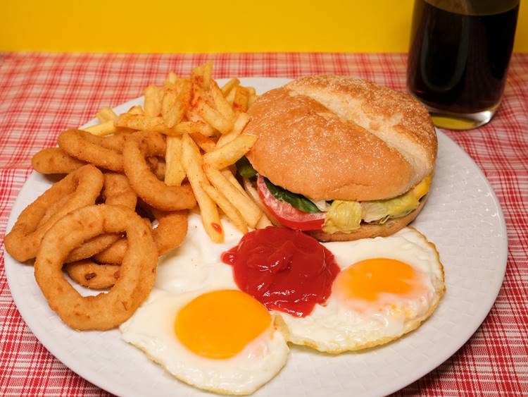 White ceramic restaurant plate with burger, onion rings, eggs and fries beside cola glass on red and white tablecloth