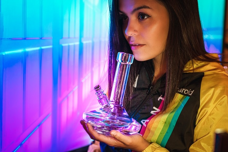 Woman holding glass bong while wearing polaroid jacket in room with neon lighting